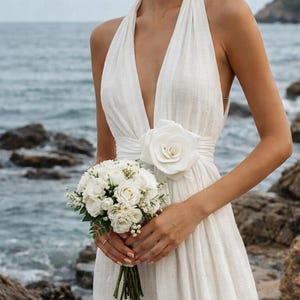 May include: A white halter-neck wedding dress with a plunging neckline and a large rose detail at the waist. The bride holds a bouquet of white roses. The background features a rocky shoreline and the ocean.