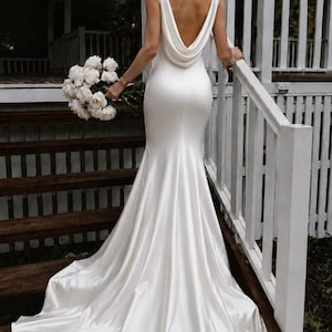 May include: A floor-length white satin gown featuring a deep cowl back, sleeveless bodice, and a fitted silhouette that flares into a train at the hem. The dress is paired with a large bouquet of white peonies held in one hand.
