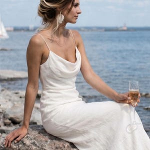 May include: A white, floor-length dress with a cowl neckline. The dress has thin straps and a flowing skirt. The person is sitting on a rock near the ocean, holding a champagne flute. The background includes the ocean and a blue sky.