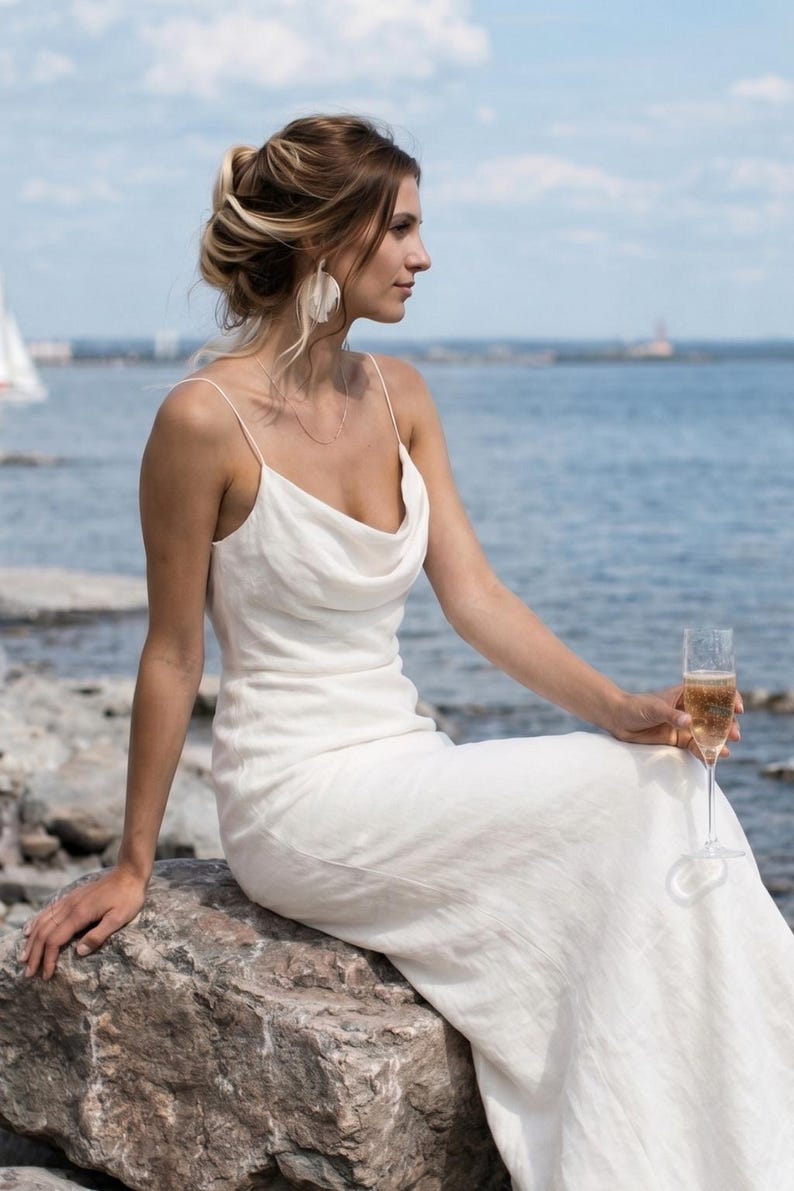 May include: A white, floor-length dress with a cowl neckline. The dress has thin straps and a flowing skirt. The person is sitting on a rock near the ocean, holding a champagne flute. The background includes the ocean and a blue sky.