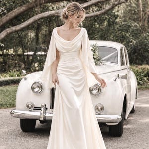May include: A floor-length ivory gown featuring a draped cowl neckline, flowing cape-style sleeves, and a fitted bodice with subtle ruching at the waist. The skirt falls in a smooth, straight silhouette to the floor, crafted from a satin-like fabric.