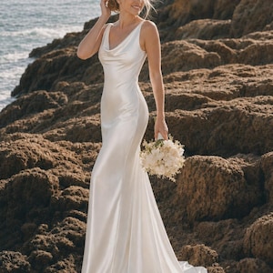 May include: A floor-length white satin gown featuring a cowl neckline, sleeveless bodice, and a fitted silhouette that flares slightly at the hem. The dress is paired with a small, round bouquet of white flowers and greenery held in one hand.