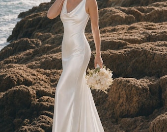 May include: A floor-length white satin gown featuring a cowl neckline, sleeveless bodice, and a fitted silhouette that flares slightly at the hem. The dress is paired with a handheld bouquet of white flowers and greenery.