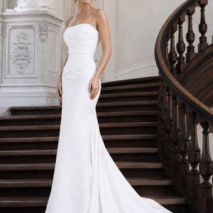 May include: A floor-length white gown featuring a straight neckline, thin spaghetti straps, and a fitted bodice with horizontal ruched detailing that transitions into a flared skirt with a train, creating a sleek and structured silhouette.