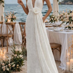 May include: A white halter-neck wedding dress with a plunging neckline and a large floral accent at the waist. The dress has a flowing skirt and is worn at an outdoor beach wedding. The bride is holding a glass of white wine.