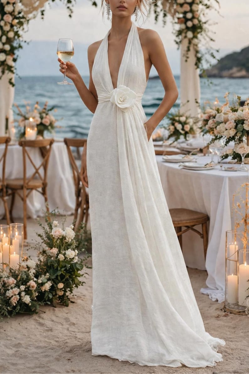 May include: A white halter-neck wedding dress with a plunging neckline and a large floral accent at the waist. The dress has a flowing skirt and is worn at an outdoor beach wedding. The bride is holding a glass of white wine.