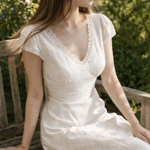 May include: A white linen dress with a V-neckline and short sleeves. The dress features delicate lace trim along the neckline and sleeves. The dress is a light, neutral color and appears to be made of a soft, breathable fabric.