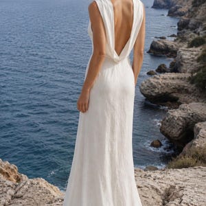 K&ouml;nnte beinhalten: Ein wei&szlig;es, &auml;rmelloses Hochzeitskleid mit tiefem, drapiertem R&uuml;cken. Das Kleid ist bodenlang und aus flie&szlig;endem Stoff gefertigt. Die Person steht auf einer felsigen Klippe mit Blick auf das Meer.