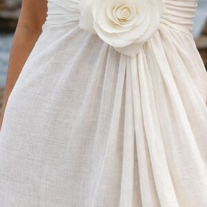 May include: Ivory-colored dress with a large, white rose accent at the waist. The dress features a draped design and a V-neckline. The fabric appears to be a textured linen or similar material, creating a soft, flowing silhouette.