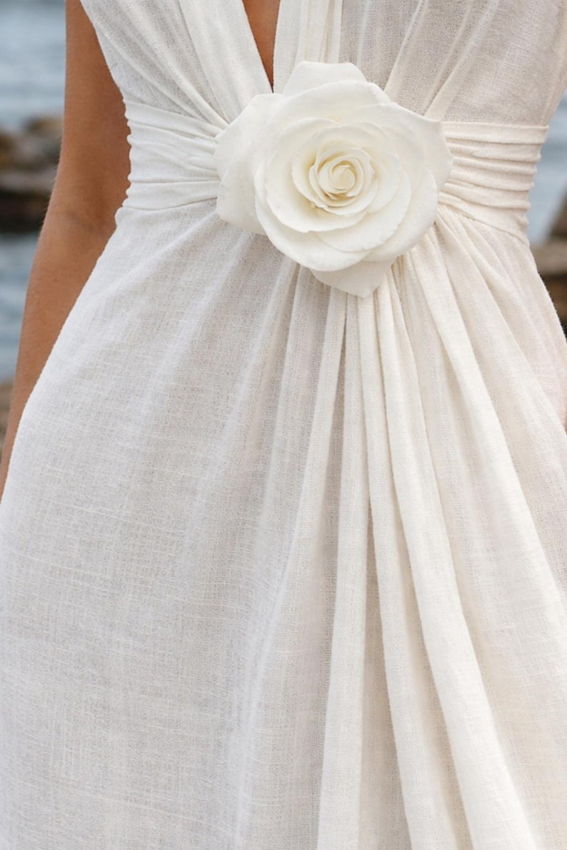 May include: Ivory-colored dress with a large, white rose accent at the waist. The dress features a draped design and a V-neckline. The fabric appears to be a textured linen or similar material, creating a soft, flowing silhouette.