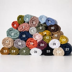 K&ouml;nnte beinhalten: Eine Sammlung gerollter Stoffe in verschiedenen Farben, darunter Blau-, Gr&uuml;n-, Rosa- und Gelbt&ouml;ne. Die Rollen sind von 1 bis 27 nummeriert und pyramidenf&ouml;rmig angeordnet. Der Stoff scheint Leinen oder ein &auml;hnliches Naturmaterial zu sein.