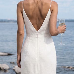 May include: A white, backless dress with spaghetti straps and a deep V-neck. The dress is made of a textured fabric and has a slim fit. A person is holding a champagne flute. The background is a body of water.