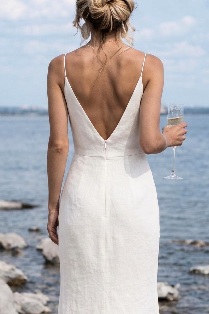 May include: A white, backless dress with spaghetti straps and a deep V-neck. The dress is made of a textured fabric and has a slim fit. A person is holding a champagne flute. The background is a body of water.