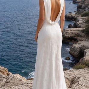 K&ouml;nnte beinhalten: Ein wei&szlig;es, &auml;rmelloses, bodenlanges Kleid mit tiefem, offenem R&uuml;cken. Das Kleid ist aus flie&szlig;endem Stoff gefertigt und wird im Freien am Meer getragen. Das Model hat blondes Haar und schaut &uuml;ber die Schulter.