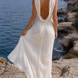 K&ouml;nnte beinhalten: Ein langes, wei&szlig;es Kleid mit tiefem R&uuml;ckenausschnitt, getragen im Freien am Meer. Das Kleid hat einen drapierten Wasserfallausschnitt und einen flie&szlig;enden Rock. Die Haare sind zu einem Dutt mit Blumen gesteckt. Das Kleid ist aus leichtem Stoff.