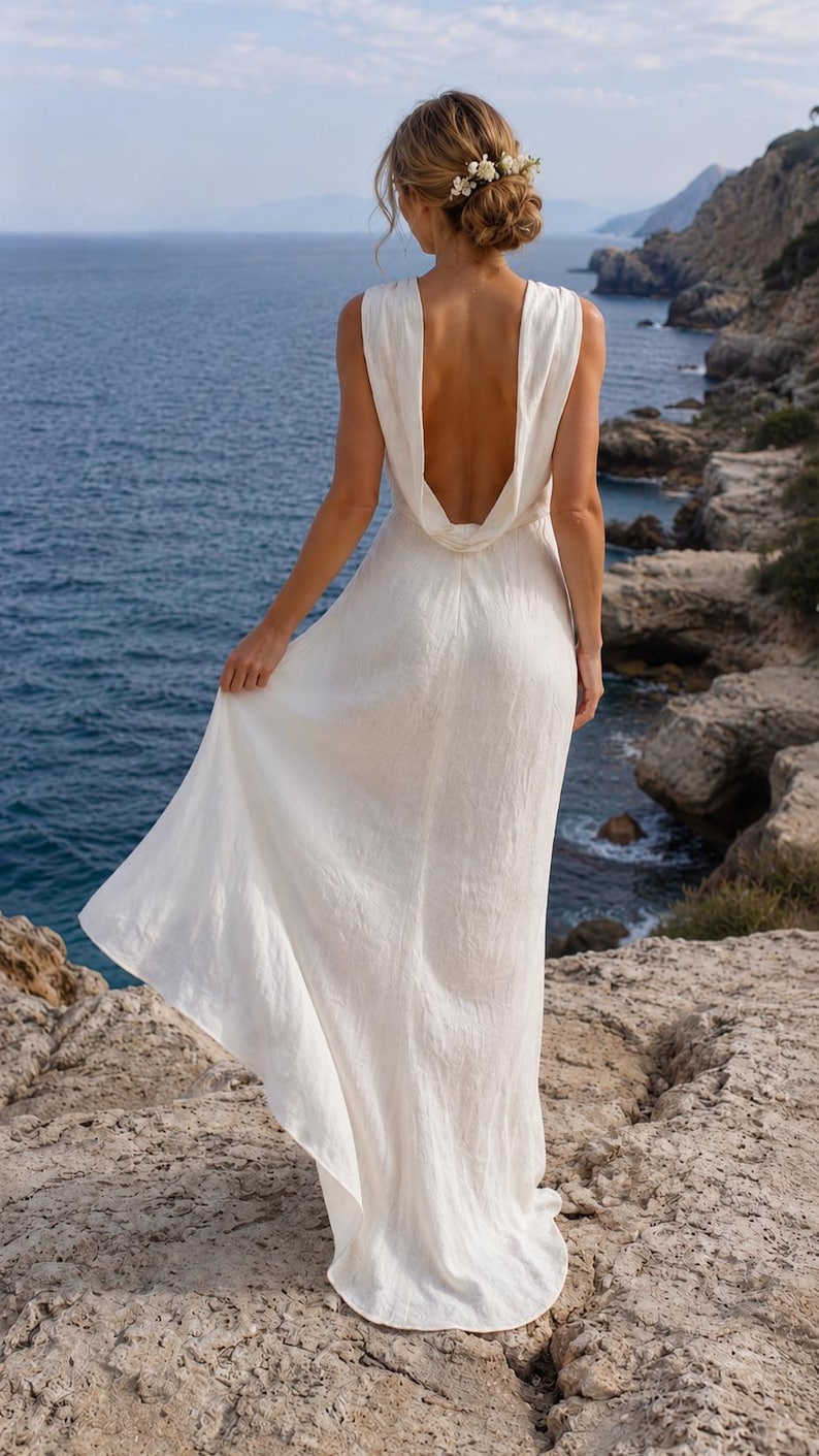 K&ouml;nnte beinhalten: Ein langes, wei&szlig;es Kleid mit tiefem R&uuml;ckenausschnitt, getragen im Freien am Meer. Das Kleid hat einen drapierten Wasserfallausschnitt und einen flie&szlig;enden Rock. Die Haare sind zu einem Dutt mit Blumen gesteckt. Das Kleid ist aus leichtem Stoff.