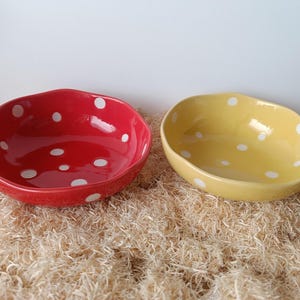 May include: Two ceramic bowls, one red and one yellow, each with a white polka dot pattern. The bowls have an irregular, wavy rim and are set on a bed of light brown material.