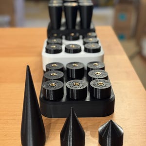 Op de afbeelding: Een verzameling zwarte 3D-geprinte objecten, waaronder cilindrische en conische vormen, en een rek met meerdere zwarte cilinders. De cilinders hebben goudkleurige accenten en tekst. De items zijn gerangschikt op een houten oppervlak.