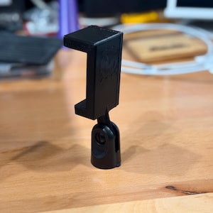 May include: A black phone holder with a pivoting base. The holder has a rectangular top and a rounded base, designed to hold a phone. The holder is made of a matte black material and sits on a wooden surface.