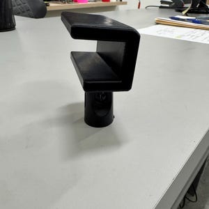 Puede incluir: Un adaptador de pie de micrófono impreso en 3D negro con una abrazadera en forma de U.