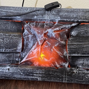 May include: A decorative electric fireplace with a faux log design. The logs are painted gray with white accents, surrounding a glowing orange and yellow flame effect. A black power cord and switch are visible.