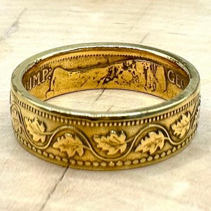 May include: A gold-toned ring crafted from a coin, featuring an exterior band embossed with a continuous pattern of stylized leaves and beaded edges. The interior surface displays remnants of original coin lettering including IMP and GEO.