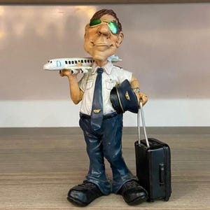 May include: A figurine of a cartoon pilot wearing a white shirt, blue tie, and blue trousers. He is holding a small aeroplane in his right hand and a black suitcase in his left hand. He is wearing sunglasses and a pilot's cap.