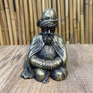 Könnte beinhalten: Eine kleine, goldfarbene Figur eines Menschen, der einen Turban und traditionelle Kleidung trägt. Die Figur sitzt mit gekreuzten Beinen und ineinander gefalteten Händen.