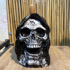 Puede incluir: Quemador de incienso de calavera negro y plateado con cabeza encapuchada. El quemador de incienso está hecho de cerámica y tiene un diseño de calavera detallado.