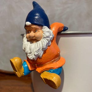 Könnte beinhalten: Eine skurrile Gartenzwergfigur mit blauem Hut, orangefarbenem Mantel und gelben Stiefeln. Der Zwerg hat einen weißen Bart und sitzt am Rand eines weißen Behälters. Ein dekorativer Artikel.