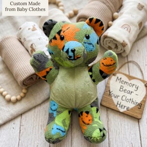 May include: A handmade green, orange, and blue teddy bear made from baby clothes. The bear has a light green belly and is surrounded by rolled-up blankets and a wooden sign that reads "Memory Bear - Your Clothing Here."