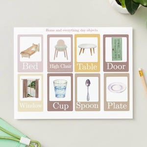 May include: A watercolor illustration of everyday objects, including a bed, high chair, table, door, window, cup, spoon, and plate. The text "Home and everything day objects" is at the top. A pencil and a green strap are in the image.