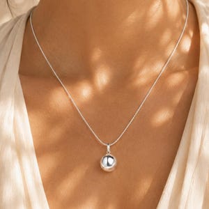 May include: A silver necklace featuring a polished, spherical ball pendant suspended from a delicate, thin silver chain. The metal surface of the round charm reflects light, creating a bright, metallic sheen against the simple, minimalist design.