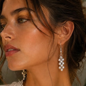 May include: Silver dangle earrings with multiple small, round beads. The earrings are hanging from a woman's ear. She is wearing a white blouse with lace trim. The background is a soft, natural green.