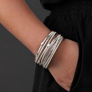 May include: A multi-strand wrap bracelet featuring several layers of light beige and tan leather cords. The design includes small, oval-shaped silver-toned metal accents and a single strand embedded with tiny, clear, sparkling crystals.
