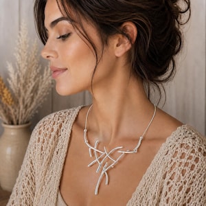 May include: A silver-toned necklace featuring a central pendant composed of multiple intersecting, smooth, curved metal bars. The pendant is attached to a thin, flexible silver-toned wire collar that rests around the neck, creating a structured look.