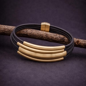 May include: A multi-strand bracelet featuring several thin black leather cords and three curved, smooth gold-toned metal bars. The bracelet is secured with a rectangular gold-toned magnetic clasp that connects the ends of the leather cords.