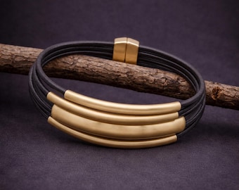 May include: A multi-strand bracelet featuring several thin black leather cords and three curved, smooth gold-toned metal bars. The bracelet is secured with a rectangular gold-toned magnetic clasp that connects the ends of the leather cords.