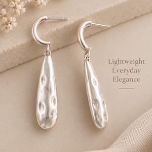 May include: A pair of silver-toned drop earrings featuring a curved post and a dangling, elongated teardrop-shaped pendant with a textured, dimpled surface. The text "Lightweight Everyday Elegance" is printed to the right of the earrings.