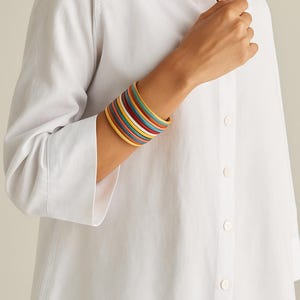 May include: A stack of thin, round bangles in a variety of colors including yellow, light blue, white, red, and dark blue. The bracelets are worn together on the wrist, creating a layered, multi-colored appearance with a smooth, uniform finish.