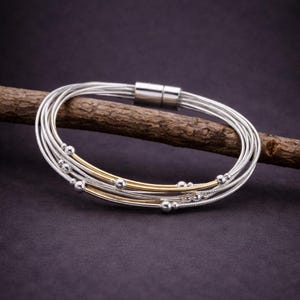 May include: A silver bracelet with multiple strands, featuring gold-coloured bands and small silver beads. The bracelet has a magnetic clasp and is displayed on a wooden branch against a dark background. Jewellery.