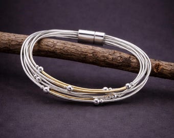 May include: A silver bracelet with multiple strands, accented with gold-colored bands and small silver beads. The bracelet has a magnetic clasp and is displayed on a wooden branch against a dark background. Jewelry.