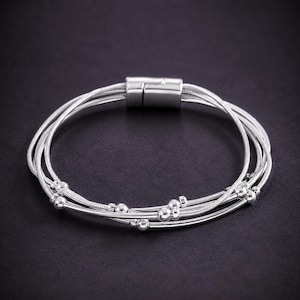 May include: A multi-strand silver-toned bracelet featuring thin snake chains and small, smooth spherical beads distributed along the strands. The bracelet is secured with a rectangular magnetic clasp that connects the multiple strands into one unit.