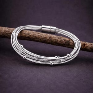 May include: A multi-strand silver bracelet featuring several thin, polished metallic chains layered together. The design includes small, round silver beads interspersed along the strands and a cylindrical magnetic clasp closure.
