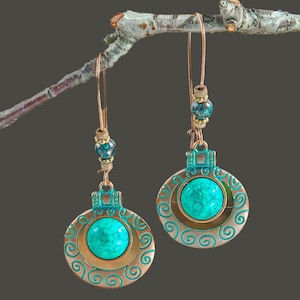Può includere: Orecchini color rame con centri in pietra turchese. Gli orecchini hanno un design circolare con un motivo a spirale decorativo e sono sospesi a un gancio di filo. Piccole perle blu e oro accentuano il design.