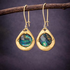 May include: A pair of gold-toned drop earrings featuring a teardrop-shaped frame with a textured finish. Each earring contains a circular, iridescent abalone shell inlay displaying vibrant shades of green, blue, and purple with a polished surface.