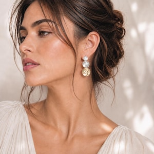 May include: A drop earring featuring three vertically aligned circular discs of varying sizes. The top disc is small and silver, the middle disc is medium-sized and silver, and the bottom disc is the largest, featuring a textured gold finish.
