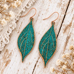 May include: A pair of drop earrings shaped like leaves with a textured, vibrant teal surface and copper-coloured edges. Each earring features a central vein detail and a curved hook for attachment, showcasing a distinct leaf-like silhouette.