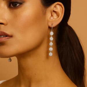 May include: Silver drop earrings featuring five graduated, sparkling, round orbs. The earrings are suspended from a small hook. The model's skin has a warm tone, and the background is a solid, light brown.