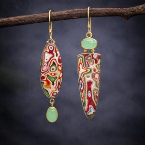 May include: A pair of gold-coloured drop earrings. One is oval with a green stone at the base, the other is a long, pointed shape with a green stone at the top. Both earrings feature a colourful, abstract pattern with red, green, and white.